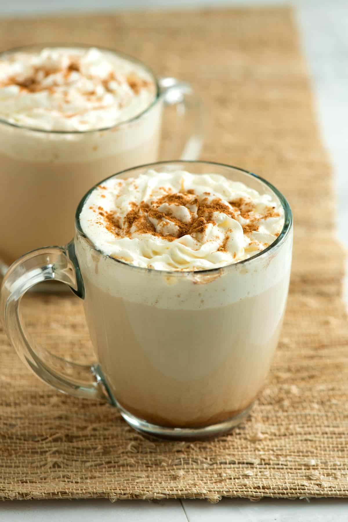 Two clear mugs are filled with pumpkin spice latte and topped with whipped cream and spices. They're placed side-by-side upon a twine-like place mat