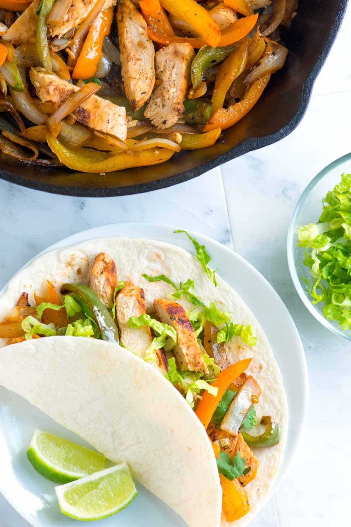 Skillet of chicken fajitas with tortillas, lettuce and limes
