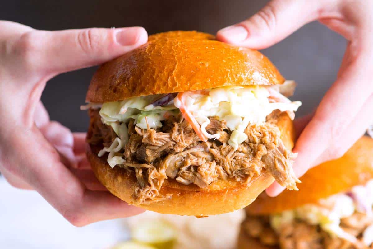 Two hands holding a sandwich filled with shredded pork and coleslaw.