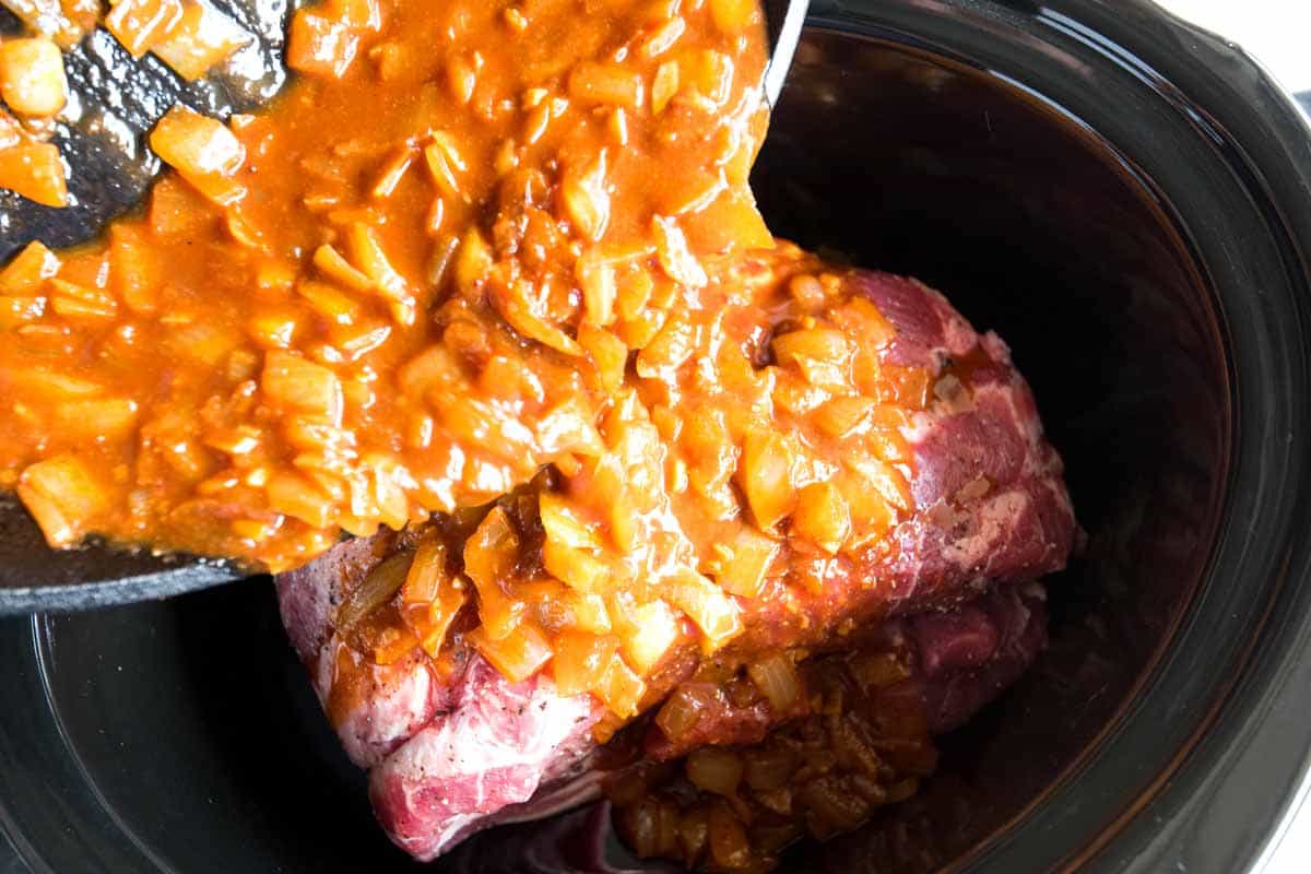 Adding onions, garlic, tomato paste, and spices on top of pork in a slow cooker for slow cooker pulled pork