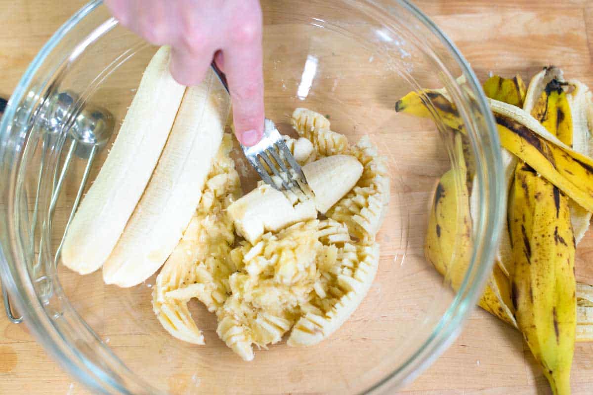 A bowl with three bananas being mashed with a fork and discarded speckled banana peel off one side.
