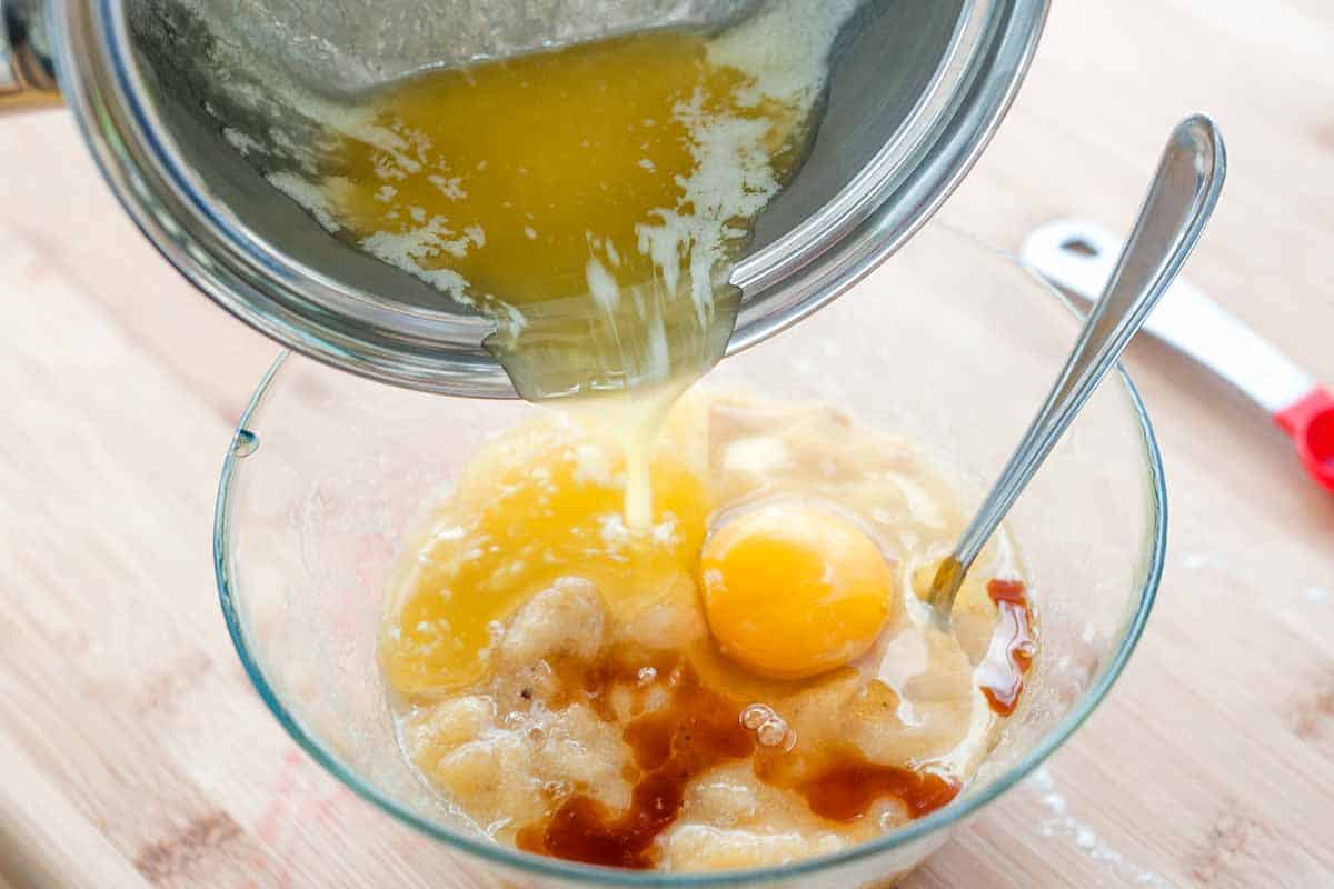 A small saucepan of melted butter is poured into a bowl with mashed bananas, an egg, and vanilla extract.