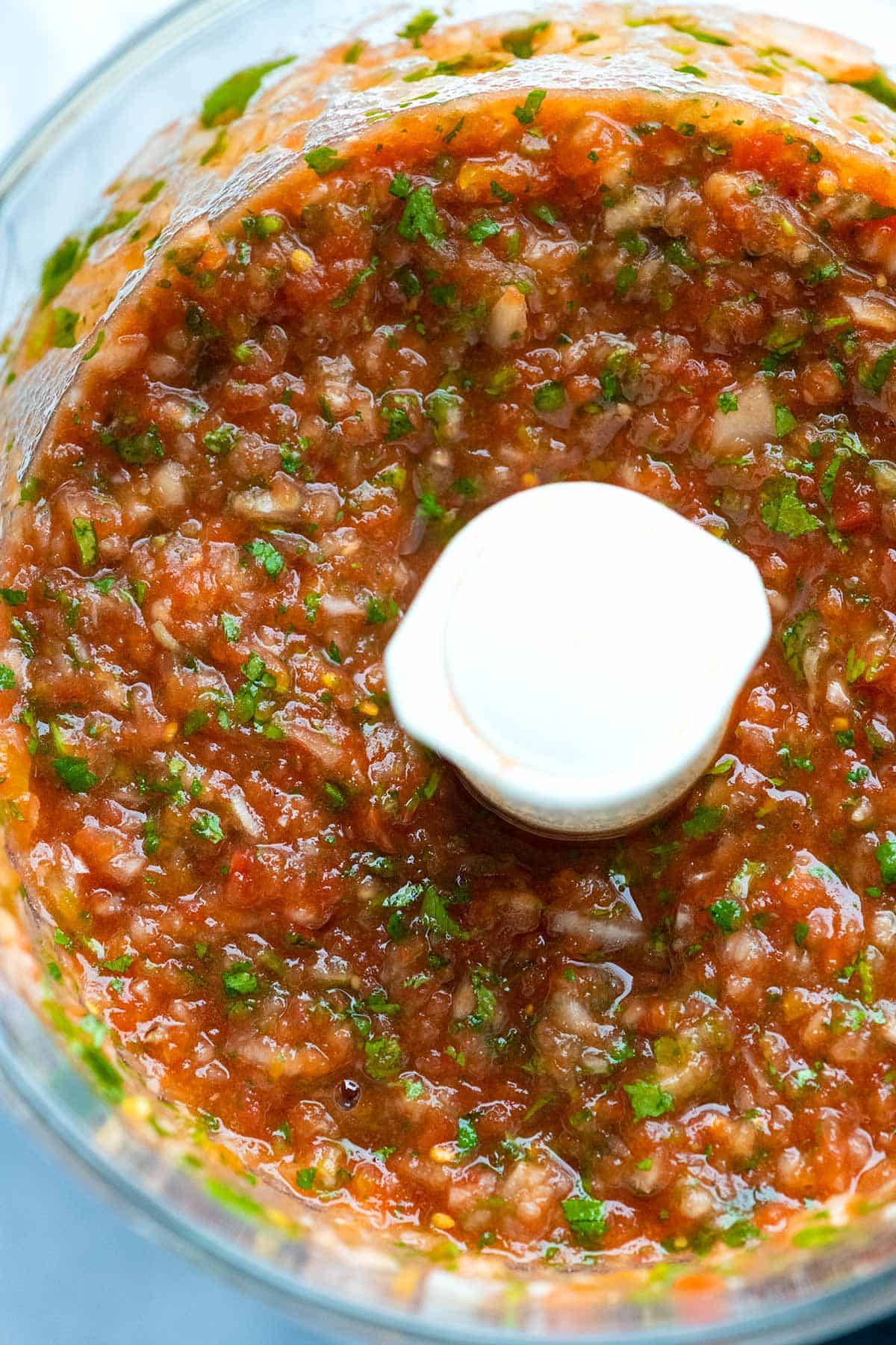 Bowl of a food processor with homemade salsa made with tomatoes, onion, jalapeño and cilantro.