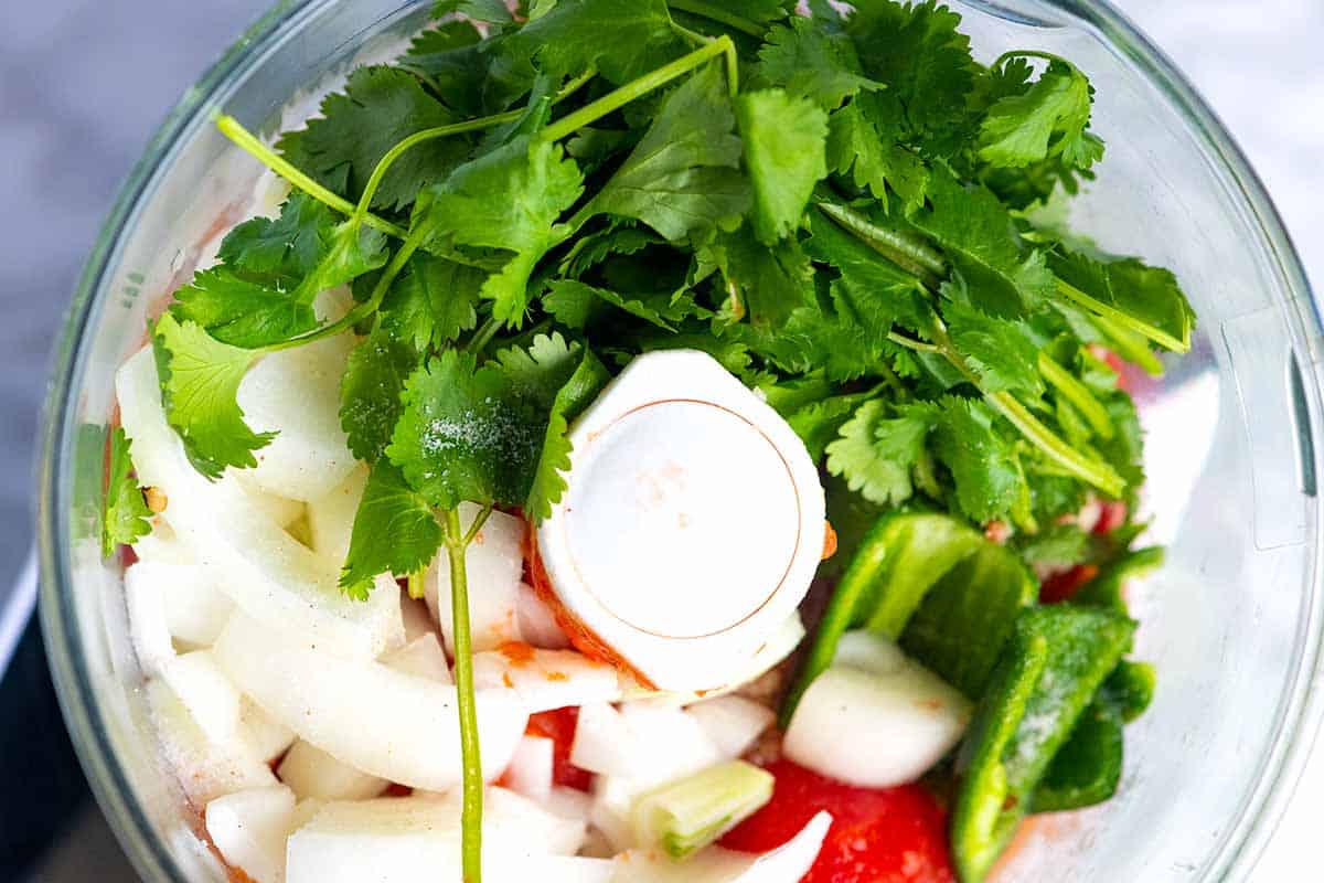 Bowl of a food processor with ingredients for salsa (tomatoes, onions, jalapeño, and cilantro).