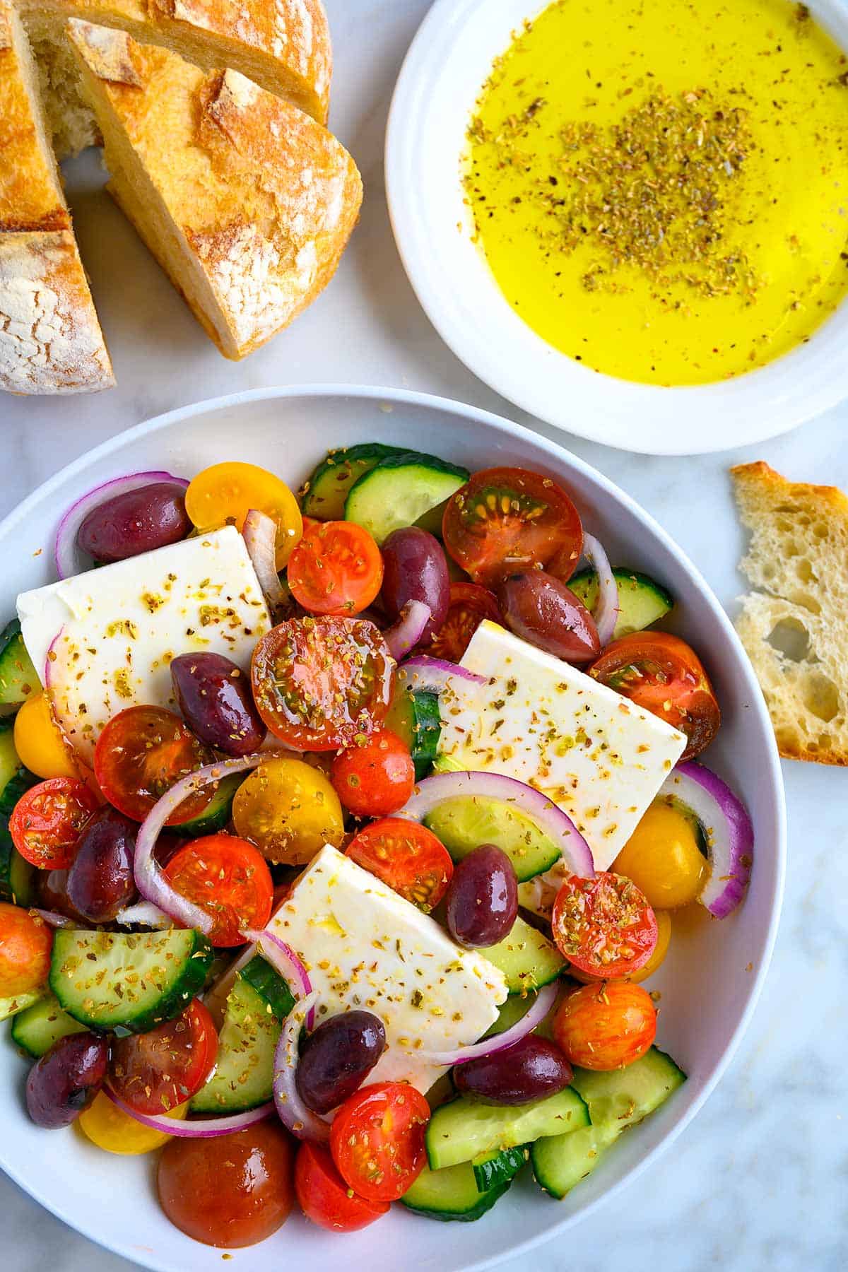 Bowl filled with a Greek salad with bread and dipping oil to the side.