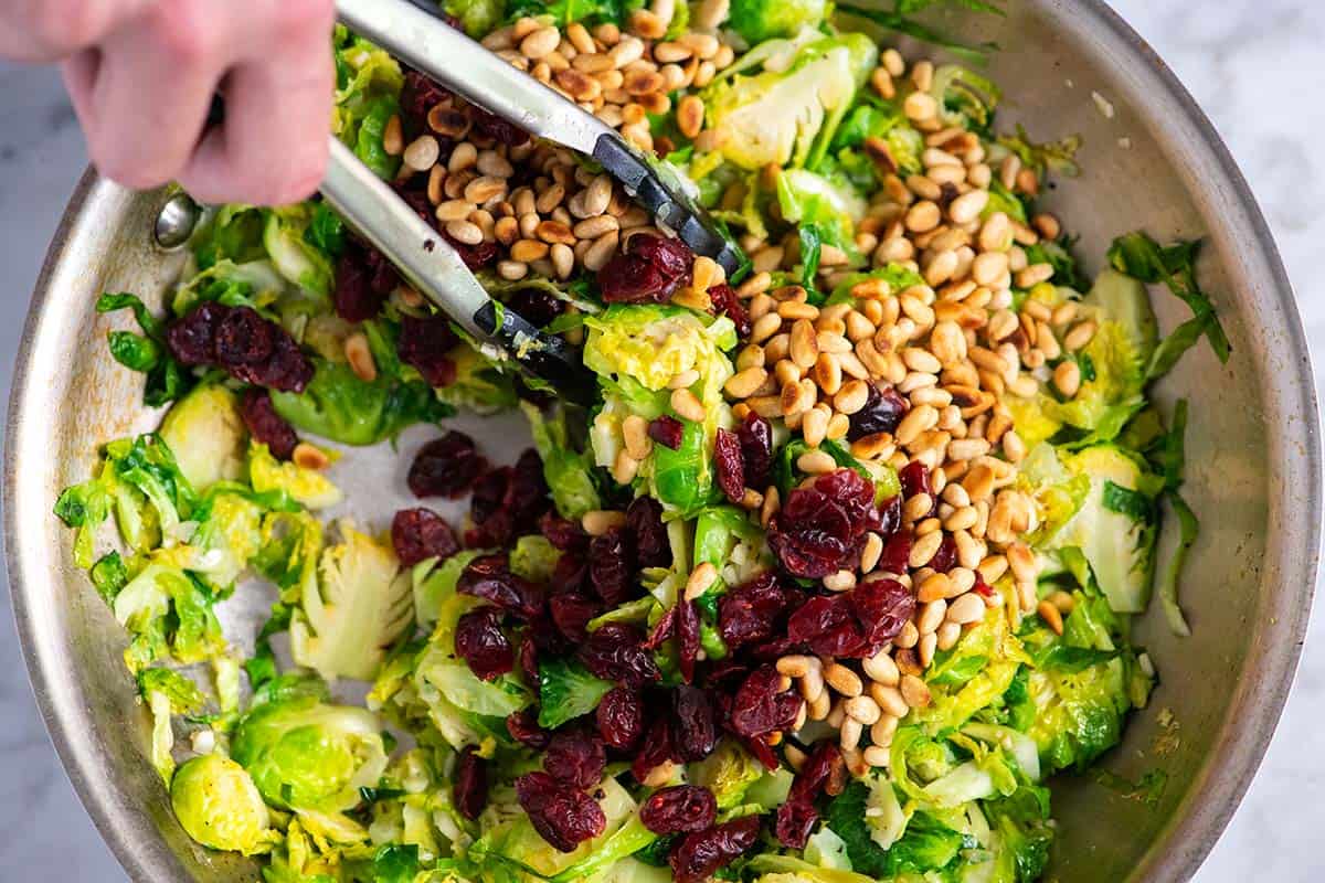 Stirring in dried cranberries and pine nuts to a skillet full of brussels sprouts