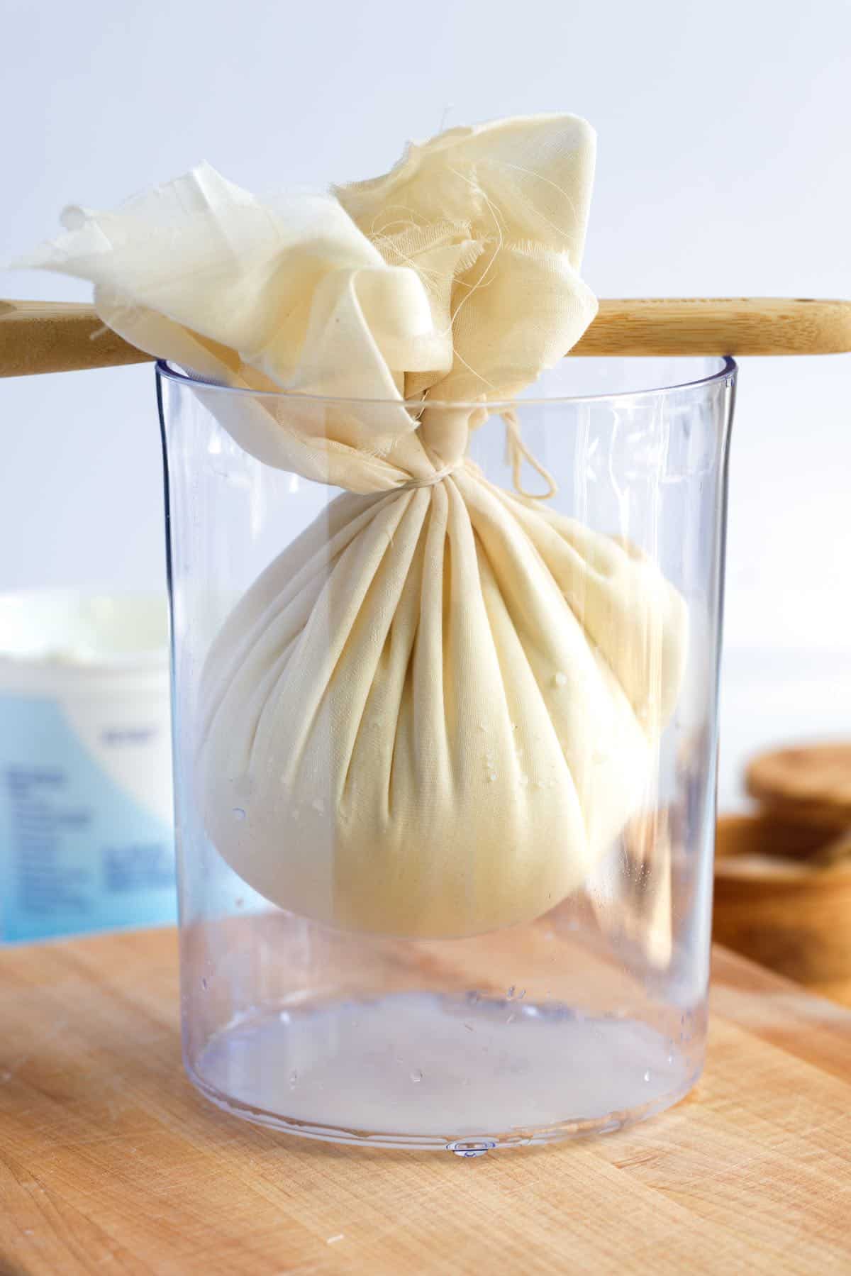 How to Make Labneh -- Cheesecloth wrapped yogurt with a wooden spoon holding it up to strain into a tall container.